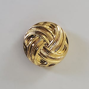 May include: A gold-toned metal button with a woven design. The button has a raised, intricate pattern that resembles a knot.