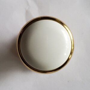 May include: A white ceramic knob with a gold rim. The knob is round and has a smooth surface.