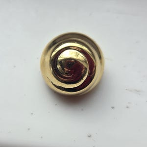 May include: A round, polished gold-tone button featuring a raised, swirling spiral pattern that radiates from the center. The surface is highly reflective, showing a smooth, metallic finish with a domed shape and a circular outer edge.