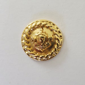 May include: A gold-toned button with a rope-like border and a raised crest in the center. The crest features a shield with an 'S' inside and a decorative design.