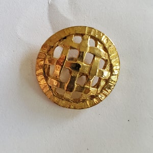 May include: A gold-colored metal button with a woven design. The button has a circular shape and a raised edge.