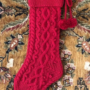May include: A vibrant red knitted Christmas stocking with a cable knit design and bobble details. The stocking has a ribbed cuff and is adorned with two small pom-poms attached by a knitted cord. The festive holiday decor is displayed on a patterned background.