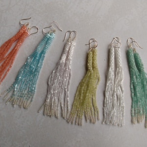 May include: Six pairs of long, beaded earrings in various colors, including peach, teal, white, green, and light green. The earrings are all made with small, round beads and have a tassel-like appearance.