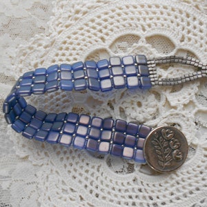 May include: A blue and silver beaded bracelet with square beads. The bracelet is made of small, square, blue beads with silver accents. A silver clasp with a floral design secures the bracelet.