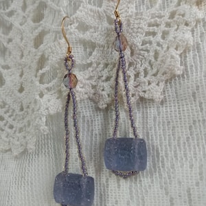 May include: A pair of long, dangling earrings with a light purple, textured, square-shaped bead at the bottom of each earring. The earrings are made with small, light purple beads and gold wire.