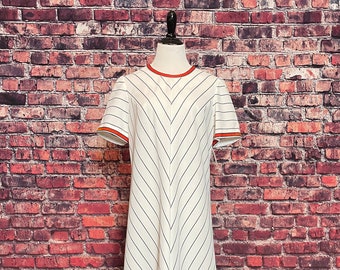 May include: A white dress with thin blue stripes and a red and blue trim around the neckline and sleeves. The dress is on a mannequin against a brick wall background.