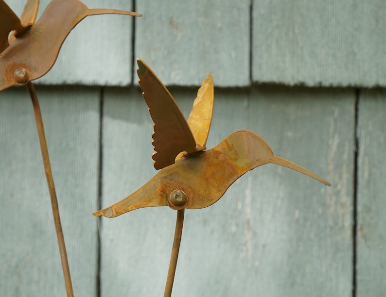 May include: Two rusty metal hummingbirds with outstretched wings, mounted on sticks against a gray wooden background.