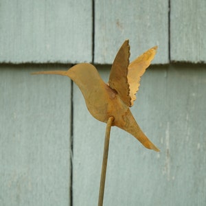 May include: A rusty metal hummingbird sculpture with outstretched wings, mounted on a thin metal rod. The sculpture is against a light blue wooden background.