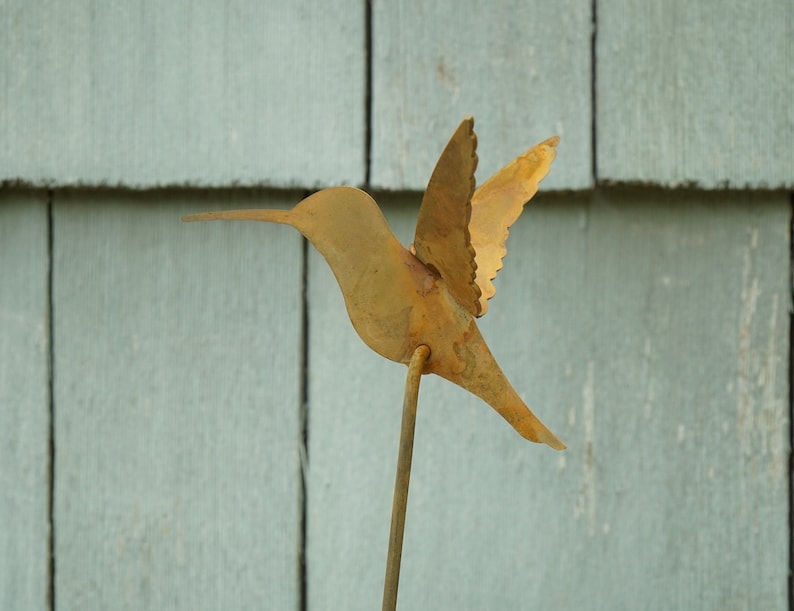 May include: A rusty metal hummingbird sculpture with outstretched wings, mounted on a thin metal rod. The sculpture is against a light blue wooden background.