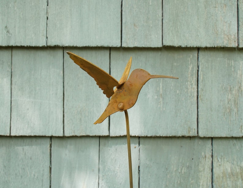 May include: A rusty metal hummingbird sculpture with wings outstretched, mounted on a metal rod. The sculpture is against a backdrop of light blue wooden siding.