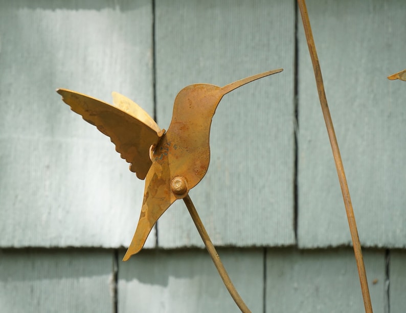 May include: A rusty metal hummingbird sculpture with outstretched wings, mounted on a metal rod.