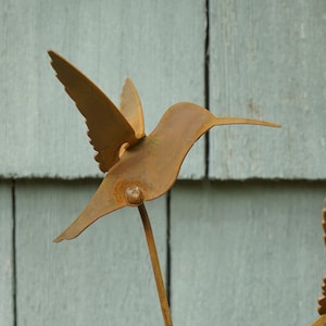 May include: A rusty metal hummingbird sculpture with outstretched wings, mounted on a metal rod. The sculpture is against a backdrop of light gray wooden siding.