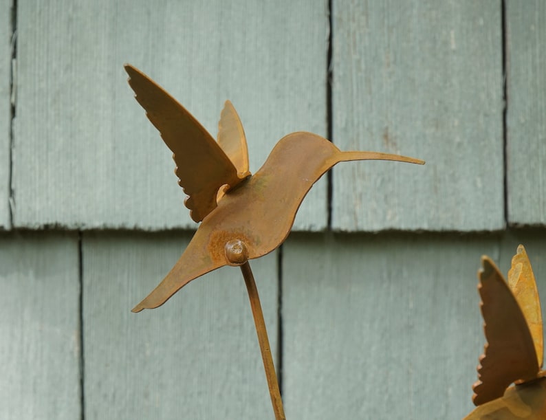 May include: A rusty metal hummingbird sculpture with outstretched wings, mounted on a metal rod. The sculpture is against a backdrop of light gray wooden siding.