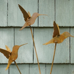May include: Three rusty metal hummingbirds with outstretched wings are mounted on thin metal rods. The hummingbirds are arranged in a triangular pattern against a light blue wooden background.