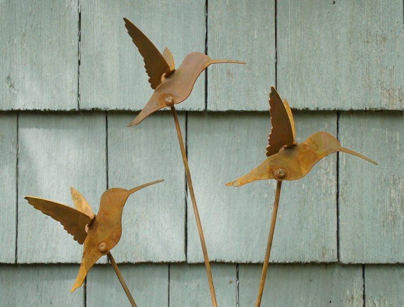 May include: Three rusty metal hummingbirds with outstretched wings are mounted on thin metal rods. The hummingbirds are arranged in a triangular pattern against a light blue wooden background.