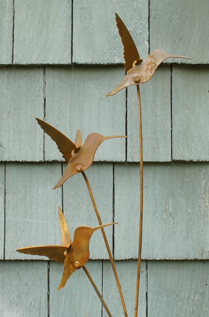 May include: Three metal hummingbirds with rusty brown wings and bodies are attached to a metal stem. The hummingbirds are arranged in a staggered formation, with the top hummingbird facing right, the middle hummingbird facing left, and the bottom hummingbird facing right. The stem is attached to a wooden surface.