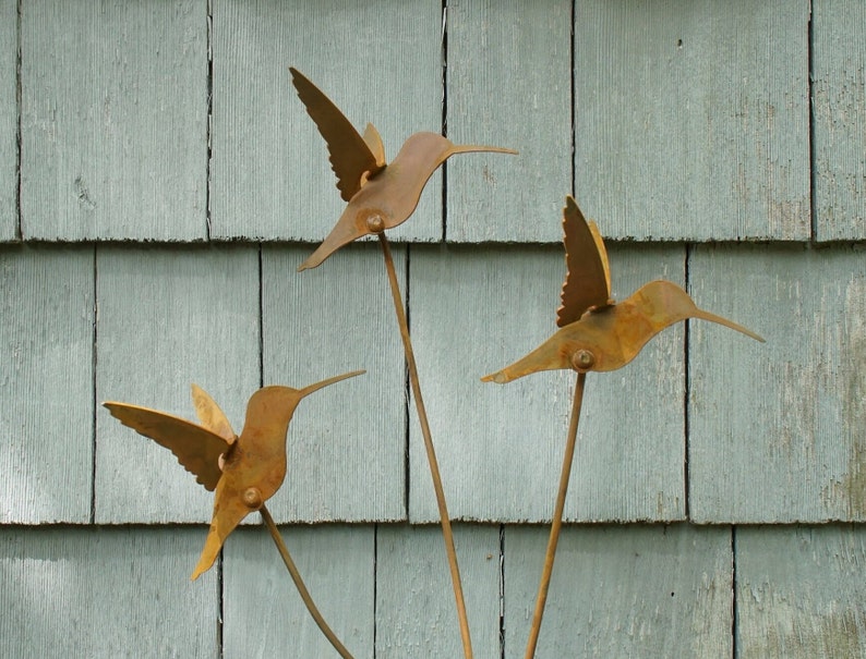 May include: Three rusty metal hummingbirds with long, thin stems are attached to a wooden surface. The hummingbirds are facing different directions and are in various positions.