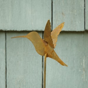 May include: A rusty metal hummingbird sculpture with a long beak and spread wings. The sculpture is mounted on a thin metal rod.