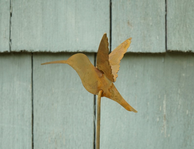 May include: A rusty metal hummingbird sculpture with a long beak and spread wings. The sculpture is mounted on a thin metal rod.