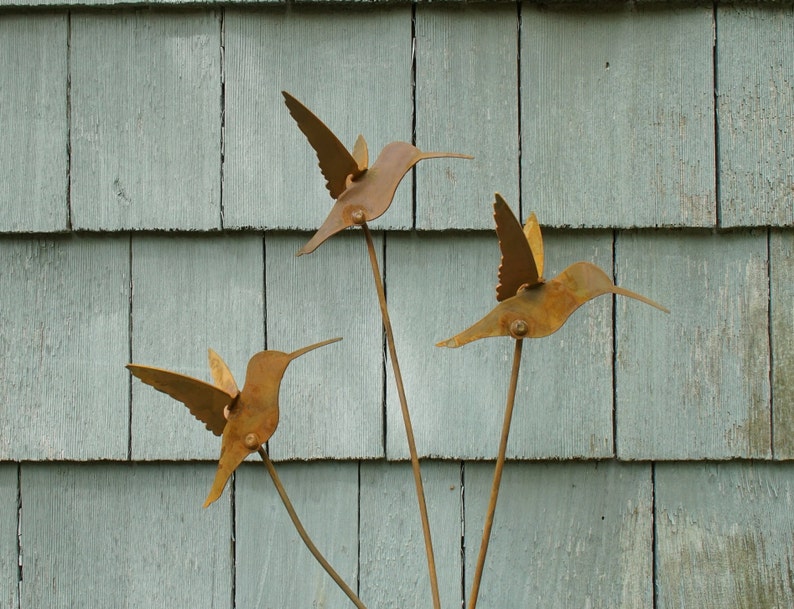 May include: Three rusty metal hummingbirds with outstretched wings are attached to metal stems. The hummingbirds are arranged in a triangular shape against a light blue wooden background.