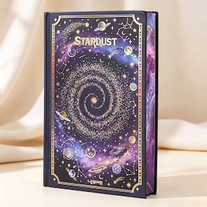 Peut inclure: Un livre à couverture rigide intitulé Stardust avec une couverture violet foncé et noire ornée d'une galaxie spirale en feuille d'or, de planètes, d'étoiles et de constellations. La tranche présente un motif nébuleuse violet et bleu.