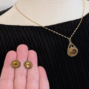 May include: A gold-toned jewelry set featuring a necklace and earrings. The necklace has a pendant with a central oval design, and the earrings are small, round, and sunburst-shaped. The jewelry is displayed on a black top.