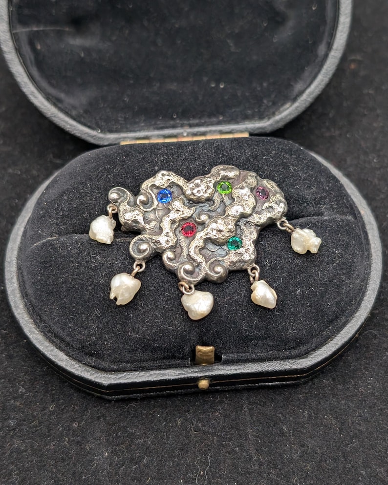 May include: A silver-toned brooch in an abstract cloud shape, adorned with colorful gemstones and dangling pearl accents. The brooch is set in a black velvet jewelry box, showcasing its intricate design and vintage appeal.