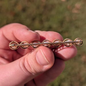 May include: A gold-toned bar pin featuring a series of small, white pearls. The pearls are set in a repeating pattern within a delicate, curved design. The pin is held against a blurred, natural background.