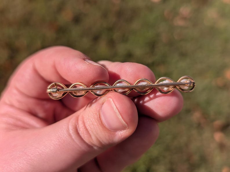 May include: A gold-toned bar pin featuring a series of small, white pearls. The pearls are set in a repeating pattern within a delicate, curved design. The pin is held against a blurred, natural background.