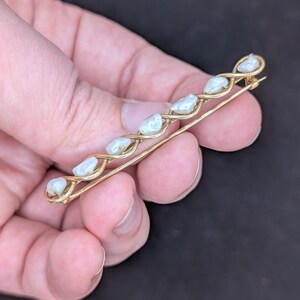 May include: A gold-toned bar pin featuring a series of white, irregularly shaped pearls. The pearls are set along a delicate, interwoven design. The pin is held in a hand against a dark background, showcasing its elegant simplicity.