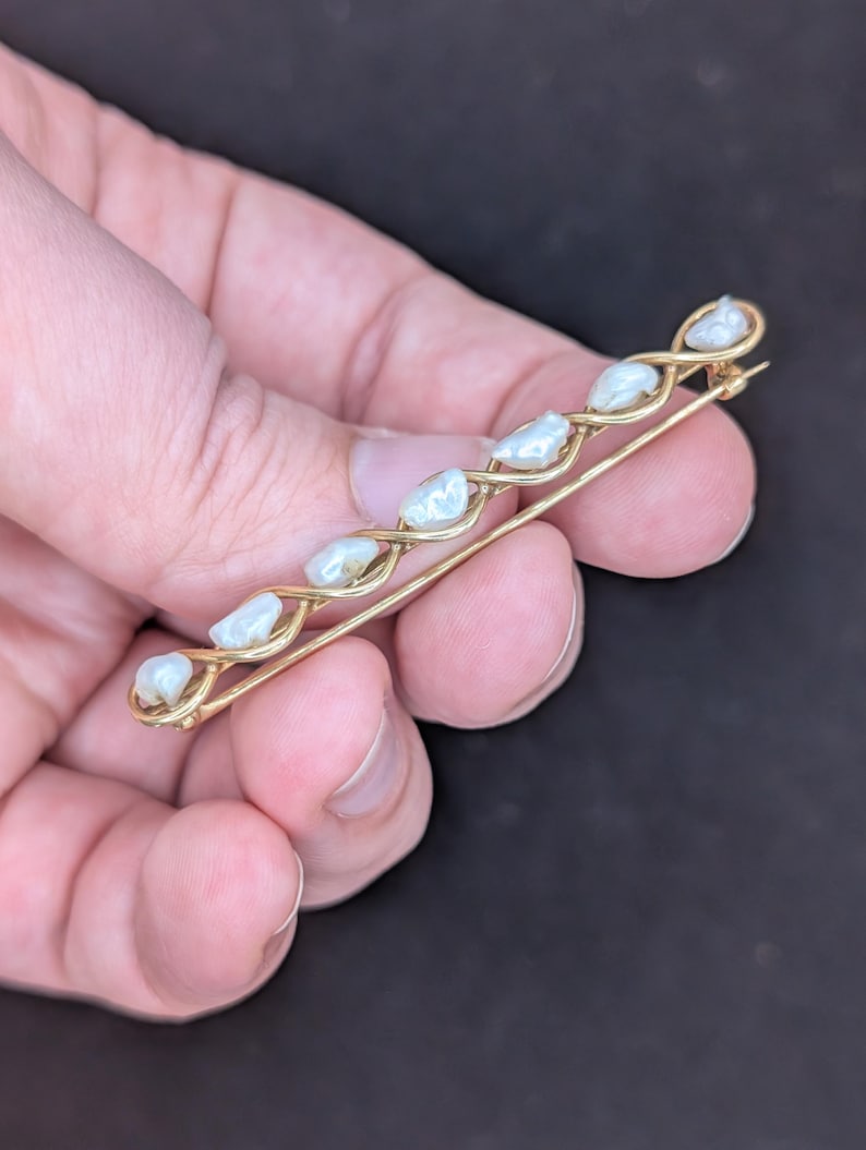 May include: A gold-toned bar pin featuring a series of white, irregularly shaped pearls. The pearls are set along a delicate, interwoven design. The pin is held in a hand against a dark background, showcasing its elegant simplicity.
