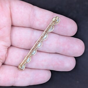 May include: A gold-toned bar pin with a series of small, evenly spaced pearls. The pin has a delicate, scalloped design along the edge where the pearls are set. The pin is held in a hand against a dark background.