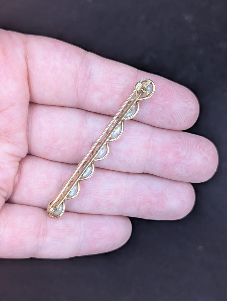 May include: A gold-toned bar pin with a series of small, evenly spaced pearls. The pin has a delicate, scalloped design along the edge where the pearls are set. The pin is held in a hand against a dark background.