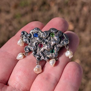 May include: A silver-toned brooch with an abstract design, adorned with colorful gemstones and dangling pearls. The brooch features a textured surface and is held in a hand, showcasing its intricate details and craftsmanship.