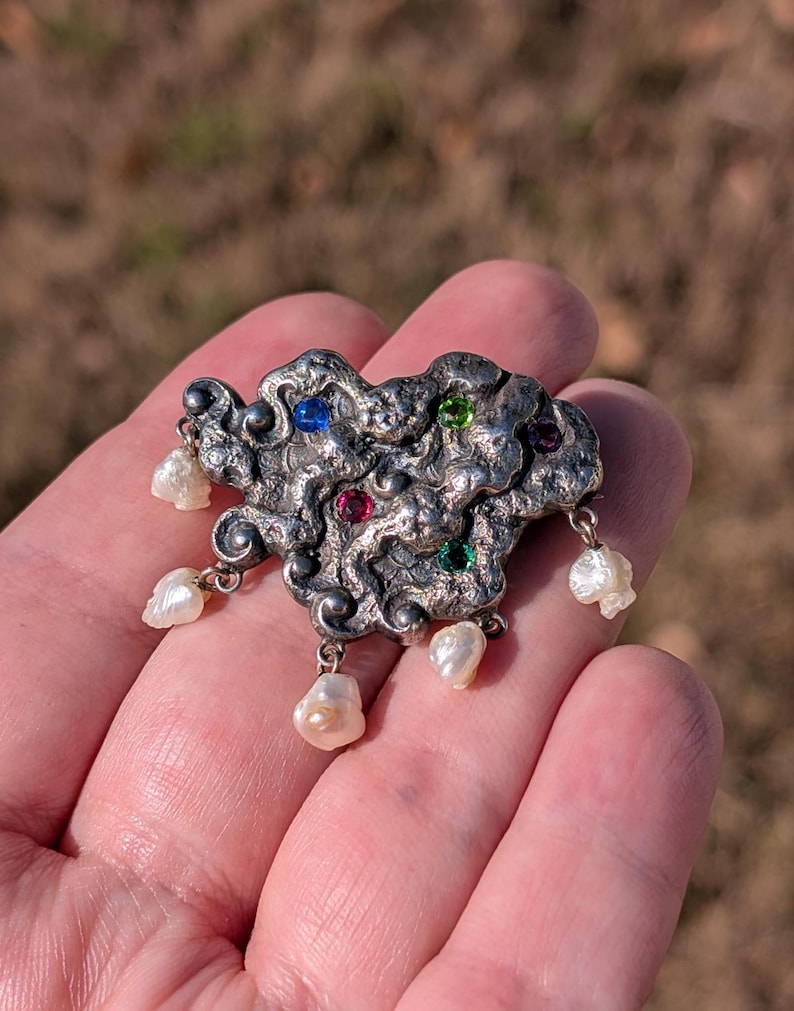 May include: A silver-toned brooch with an abstract design, adorned with colorful gemstones and dangling pearls. The brooch features a textured surface and is held in a hand, showcasing its intricate details and craftsmanship.
