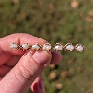 May include: A gold-toned barrette featuring a series of white pearls. The barrette has an intricate, interwoven design. The pearls are evenly spaced along the barrette, creating a delicate and elegant look.