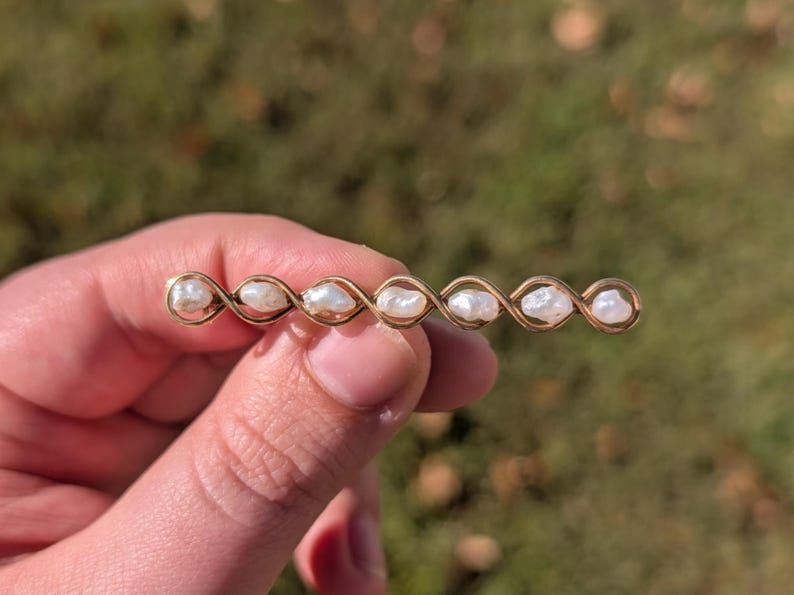 May include: A gold-toned barrette featuring a series of white pearls. The barrette has an intricate, interwoven design. The pearls are evenly spaced along the barrette, creating a delicate and elegant look.