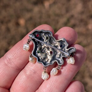 May include: Silver-toned brooch with an irregular shape, adorned with colorful gemstones and dangling pearls. The brooch features red, green, and blue stones. The pearls are teardrop-shaped and hang from the bottom of the brooch.