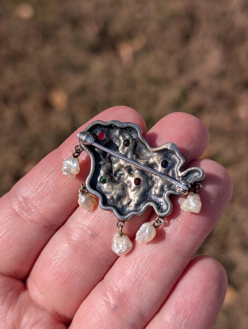May include: Silver-toned brooch with an irregular shape, adorned with colorful gemstones and dangling pearls. The brooch features red, green, and blue stones. The pearls are teardrop-shaped and hang from the bottom of the brooch.