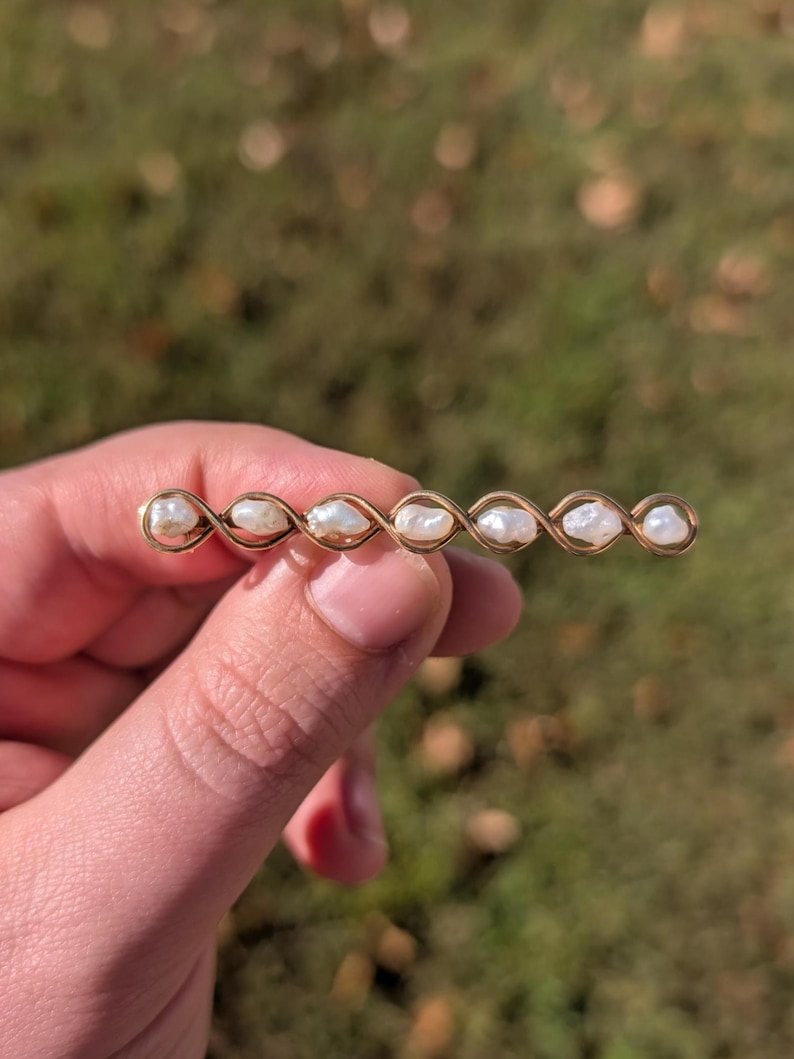 May include: A gold-toned barrette featuring six irregularly shaped pearls. The pearls are set in a twisted gold wire design. The barrette is held against a blurred green and brown background, suggesting an outdoor setting.