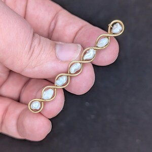 May include: A gold-toned bar pin featuring a series of five irregularly shaped, off-white pearls. The pearls are set within a delicate, interwoven gold wire design. The pin is held in a hand against a dark background.