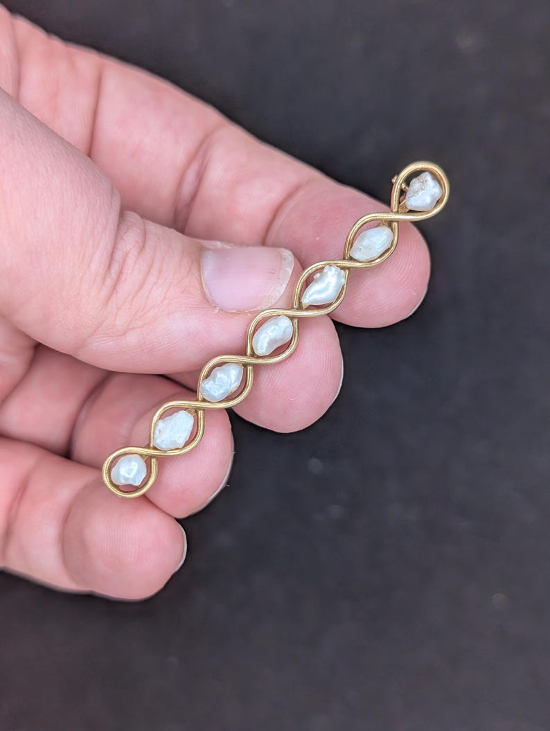 May include: A gold-toned bar pin featuring a series of five irregularly shaped, off-white pearls. The pearls are set within a delicate, interwoven gold wire design. The pin is held in a hand against a dark background.