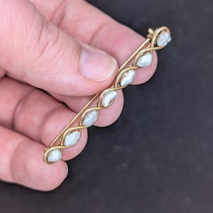 May include: A vintage gold-tone brooch featuring a series of six white, oval-shaped pearls. The pearls are set in a twisted, vine-like design. The pin has a simple clasp on the back.