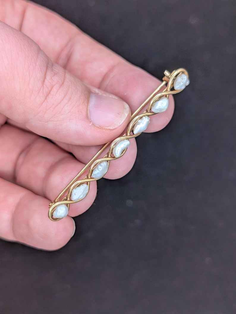 May include: A vintage gold-tone brooch featuring a series of six white, oval-shaped pearls. The pearls are set in a twisted, vine-like design. The pin has a simple clasp on the back.