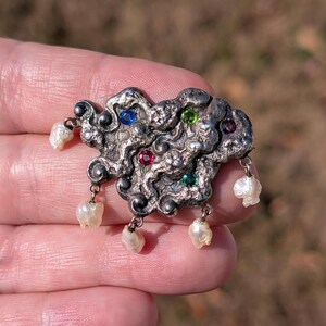 May include: A silver-toned brooch with an abstract design, featuring embedded blue, green, and red gemstones. The brooch has dangling white pearls. The background is blurred, suggesting an outdoor setting.