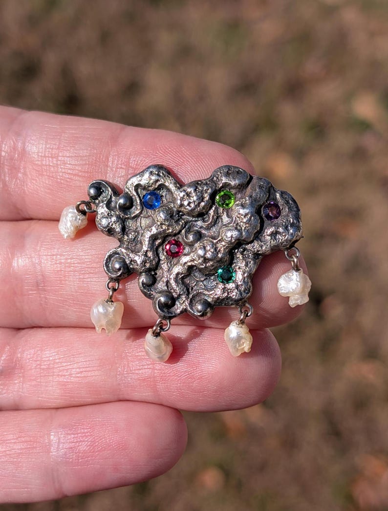 May include: A silver-toned brooch with an abstract design, featuring embedded blue, green, and red gemstones. The brooch has dangling white pearls. The background is blurred, suggesting an outdoor setting.