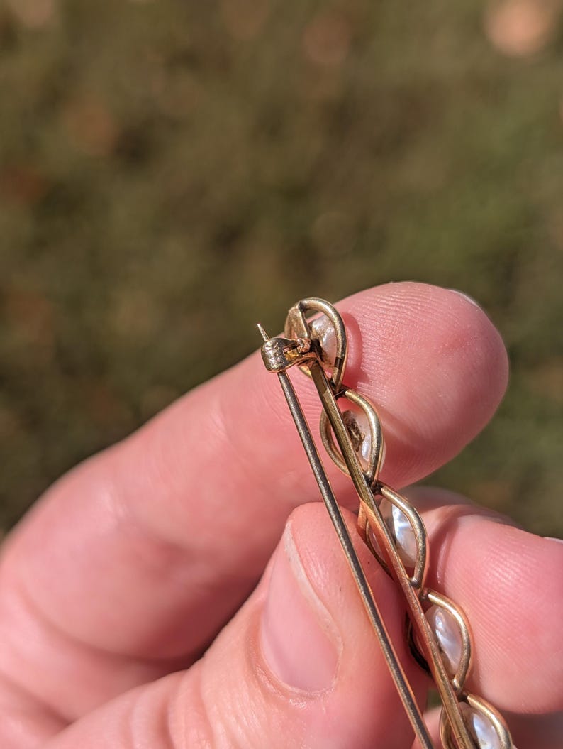 May include: Close-up of a gold-toned brooch featuring a series of oval-shaped, off-white stones. The brooch has a pin closure on the back. The background is blurred, with hints of green and brown.