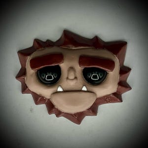 May include: A whimsical, handmade, face-shaped ornament. The ornament features a light brown face with large, black eyes, two small white fangs, and a red, spiky border. The overall design is cartoonish and expressive, with a slightly sad or contemplative expression.