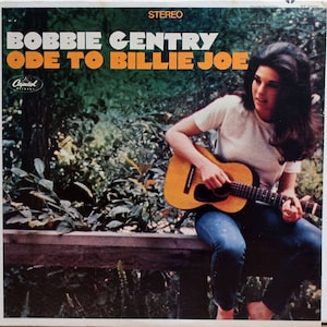 Op de afbeelding: Een vrouw zit op een houten hek en speelt een bruine akoestische gitaar. Ze draagt een wit T-shirt en een blauwe spijkerbroek. De albumhoes heeft de tekst "Bobbie Gentry Ode To Billie Joe" met een Capitol Records-logo.