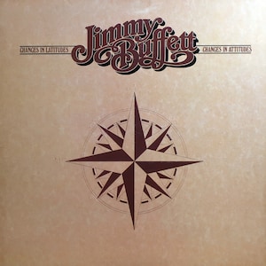 Pode incluir: Capa do álbum com o nome "Jimmy Buffett" em uma fonte marrom estilizada acima de um desenho de rosa dos ventos. O título do álbum "Changes in Latitudes, Changes in Attitudes" é impresso em texto marrom sobre um fundo bege.